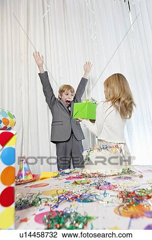 Stock Image - Sister giving gift to surprised birthday boy with arms raised.. Fotosearch
