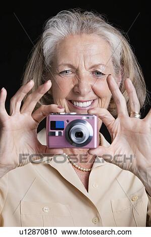Stock Image - Smiling senior woman taking picture with camera against black background. Fotosearch