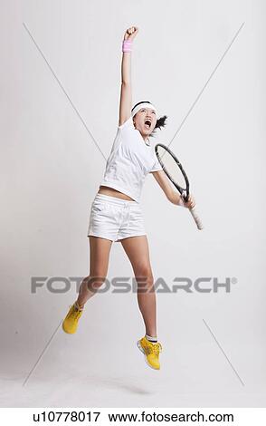 Stock Photo - Young Asian female tennis player with racket jumping in excitement against white background. Fotosearch