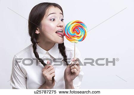 Stock Photography - Young Asian woman licking a multicolored lollipop against white background. Fotosearch
