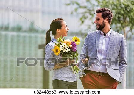 Stock Image - Young attractive business couple walking in park while the woman is holding boquet of flowers. Fotosearch