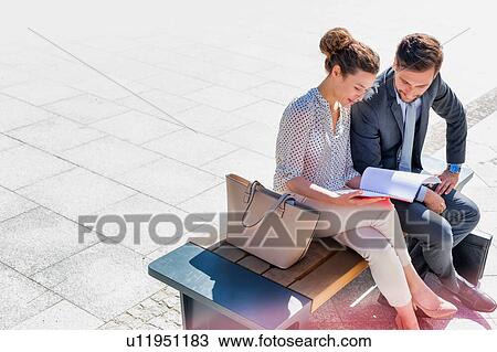 Stock Image - Young attractive businessman discussing business plans over document with beautiful businesswoman while sitting on bench against office building. Fotosearch