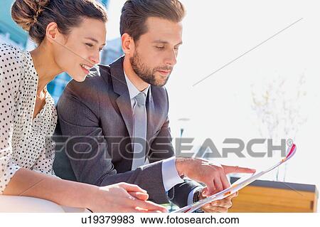 Stock Image - Young attractive businessman discussing business plans over document with beautiful businesswoman while sitting on bench against office building. Fotosearch