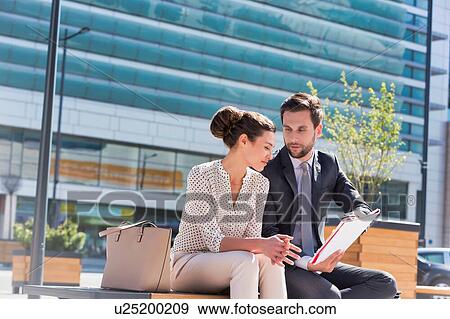 Stock Photo - Young attractive businessman discussing business plans over document with beautiful businesswoman while sitting on bench against office building. Fotosearch