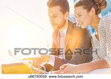 Stock Photo - Young attractive businessman discussing business plans over document with beautiful businesswoman while sitting on bench against office building. Fotosearch