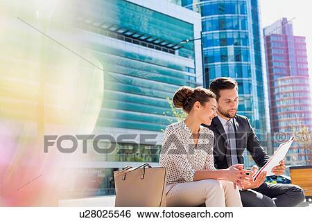 Young attractive businessman discussing business plans over document with beautiful businesswoman while sitting on bench against office building View Large Photo Image Stock Photograph - Young attractive businessman discussing business plans over document with beautiful businesswoman while sitting on bench against office building. Fotosearch