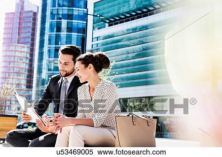 Stock Photography - Young attractive businessman discussing business plans over document with beautiful businesswoman while sitting on bench against office building. Fotosearch