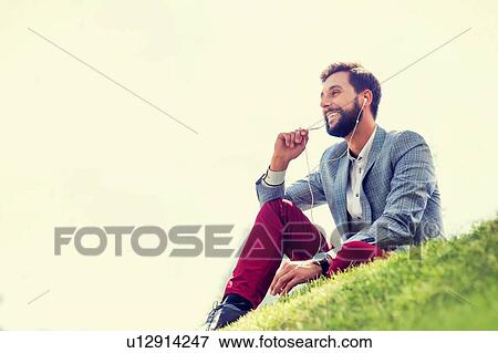 Stock Photo - Young attractive businessman talking on smartphone with earphones on while sitting on the grass. Fotosearch