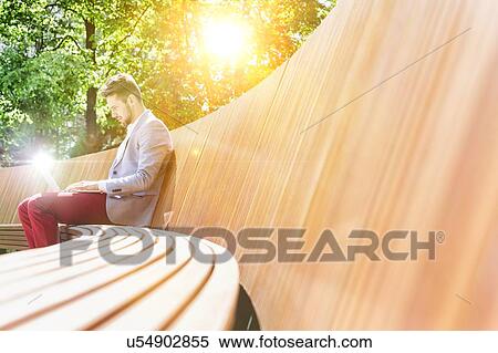 Stock Photography - Young attractive businessman working on his laptop while sitting on bench. Fotosearch