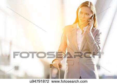 Stock Image - Young attractive businesswoman talking on smartphone while walking with her suitcase in airport. Fotosearch