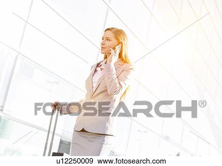 Stock Photo - Young attractive businesswoman talking on smartphone while walking with her suitcase in airport. Fotosearch