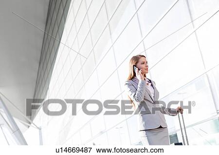 Stock Photo - Young attractive businesswoman talking on smartphone while walking with her suitcase in airport. Fotosearch