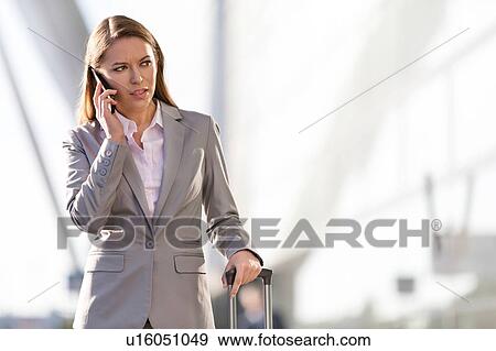 Stock Photo - Young attractive businesswoman talking on smartphone while walking with her suitcase in airport. Fotosearch