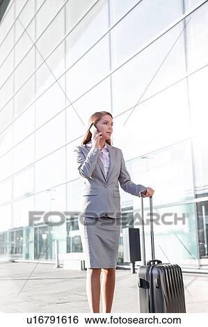 Stock Photograph - Young attractive businesswoman talking on smartphone while walking with her suitcase in airport. Fotosearch