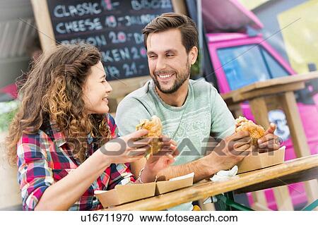 Stock Image - Young attractive couple eating hamburgers against food truck. Fotosearch