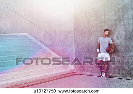 Stock Image - Young attractive man leaning on wall while holding ball. Fotosearch