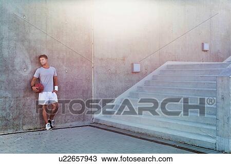 Stock Image - Young attractive man leaning on wall while holding ball. Fotosearch