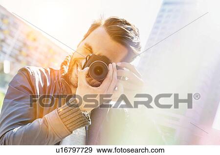 Stock Photo - Young attractive man photographing the beautiful views in the city with lens flare in background. Fotosearch