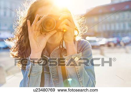 Stock Photo - Young attractive woman holding camera and taking photo with lens flare in background. Fotosearch