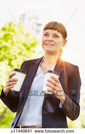 Stock Image - Young beautiful businesswoman holding two cups of coffee with lens flare in background. Fotosearch