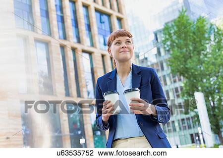 Stock Image - Young beautiful businesswoman holding two cups of coffee with lens flare in background. Fotosearch