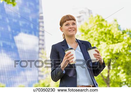 Stock Image - Young beautiful businesswoman holding two cups of coffee with lens flare in background. Fotosearch