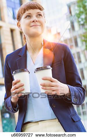 Stock Photo - Young beautiful businesswoman holding two cups of coffee with lens flare in background. Fotosearch
