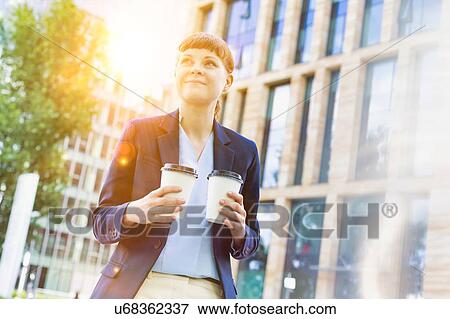 Stock Photo - Young beautiful businesswoman holding two cups of coffee with lens flare in background. Fotosearch