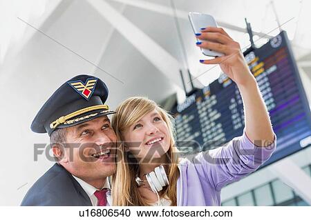 Stock Image - Young beautiful teenage girl taking selfie with mature pilot in airport. Fotosearch