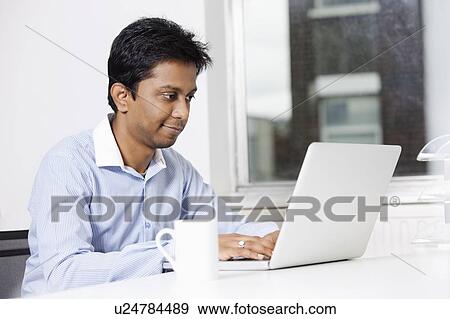 Stock Photo - Young businessman using laptop at desk in office. Fotosearch