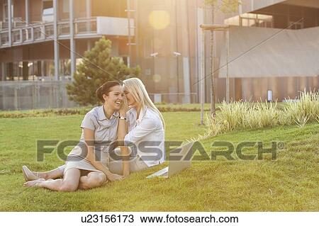 Stock Image - Young businesswoman whispering to female colleague on office lawn. Fotosearch