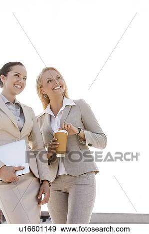 Stock Photo - Young businesswomen with disposable cup and laptop looking away outdoors. Fotosearch