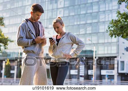 Stock Photo - Young couple standing while using smartphone after working out. Fotosearch