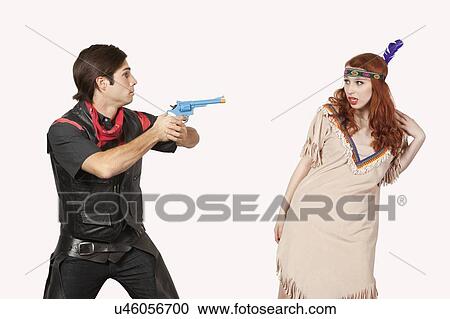 Stock Image - Young cowboy aiming gun at woman in old-fashioned costume against gray background. Fotosearch