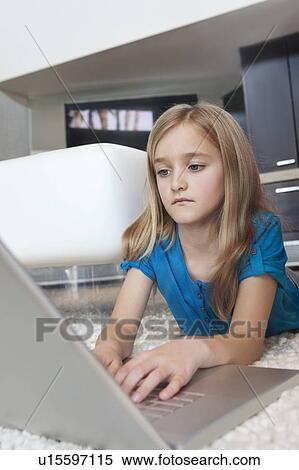 Stock Photography - Young girl using laptop while lying on rug in house. Fotosearch