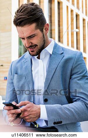 Stock Image - Young handsome businessman using smartphone against office building. Fotosearch
