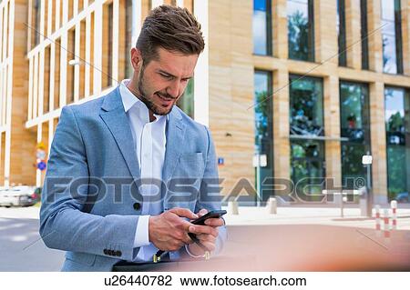 Stock Image - Young handsome businessman using smartphone against office building. Fotosearch