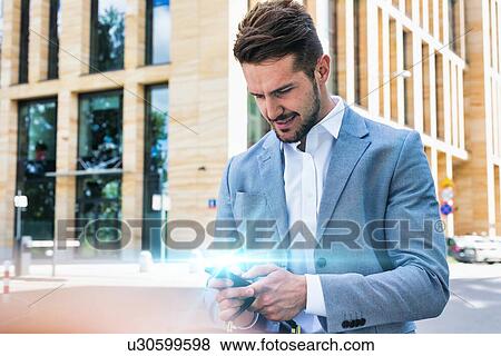 Stock Photo - Young handsome businessman using smartphone against office building. Fotosearch
