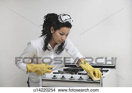 Picture - Young maidservant cleaning stove against gray background. Fotosearch