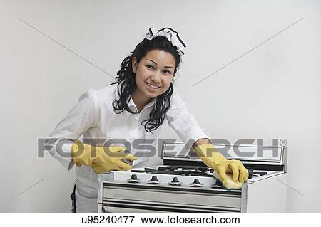 Stock Photo - Young maidservant looking away while cleaning stove against gray background. Fotosearch