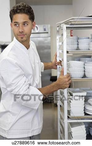 Stock Photograph - Young male chef by rack of utensils. Fotosearch