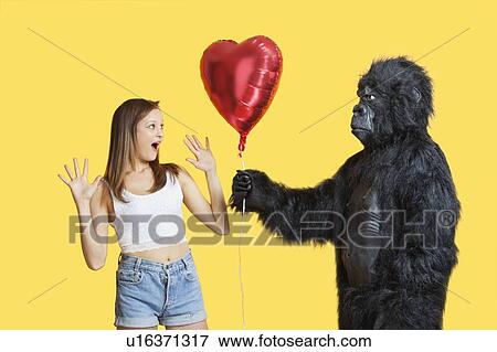 Stock Photo - Young man dressed in gorilla costume gifting heart shaped balloon to surprised woman. Fotosearch