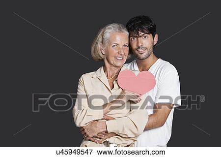 Stock Photo - Young man embracing senior woman holding red paper heart against black background. Fotosearch