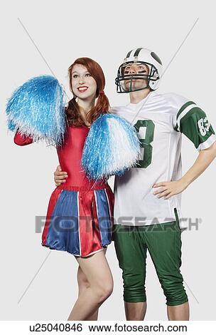 Stock Photograph - Young man in football uniform and cheerleader with pom-pom standing over gray background. Fotosearch