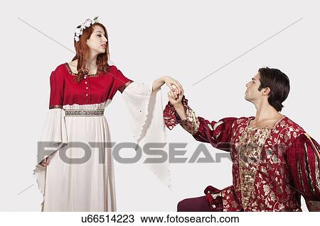 Stock Image - Young man in old-fashioned costumes holding princess's hand against gray background. Fotosearch