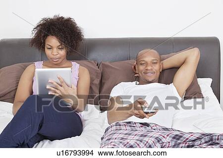 Picture - Young man watching TV while woman using tablet PC in bed. Fotosearch