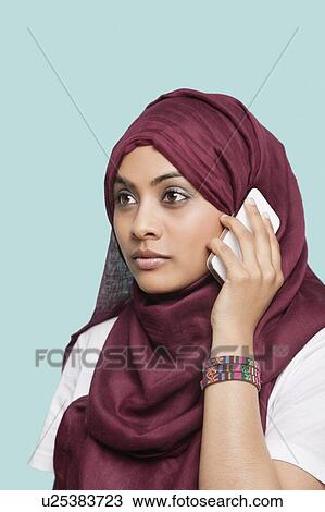 Stock Image - Young Muslim woman in maroon headcloth using cell phone against blue background. Fotosearch