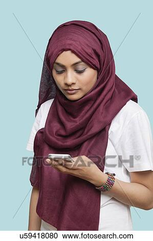 Stock Image - Young Muslim woman in traditional clothing text messaging against blue background. Fotosearch