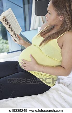 Stock Photo - Young pregnant woman reading book while relaxing on bed at home. Fotosearch