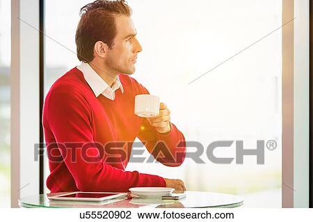 Stock Image - Young thoughtful businessman sitting while drinking coffee in lobby with lens flare. Fotosearch